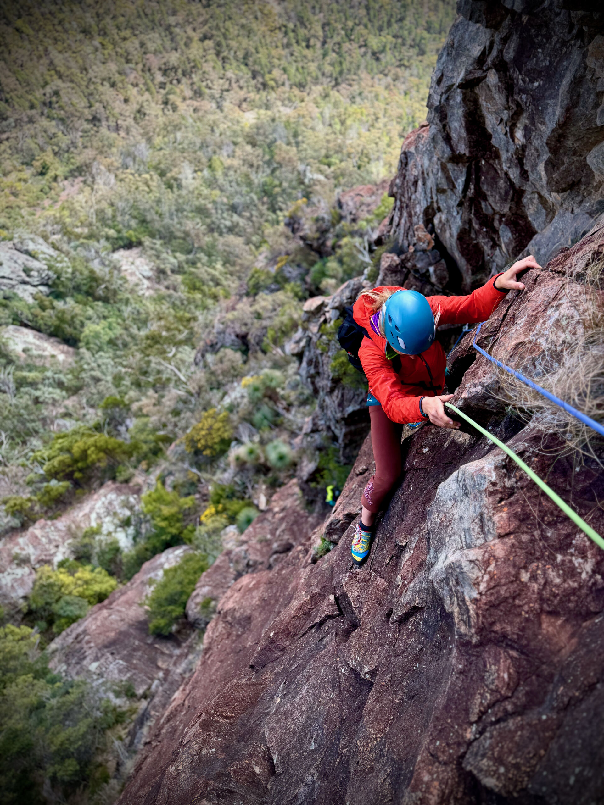 A climber with a bright red jacket and blue helmet climbs toward the camera. She is looking down at her feet trying to make the best use of them. The background is a long way down, showing how high the climber is.