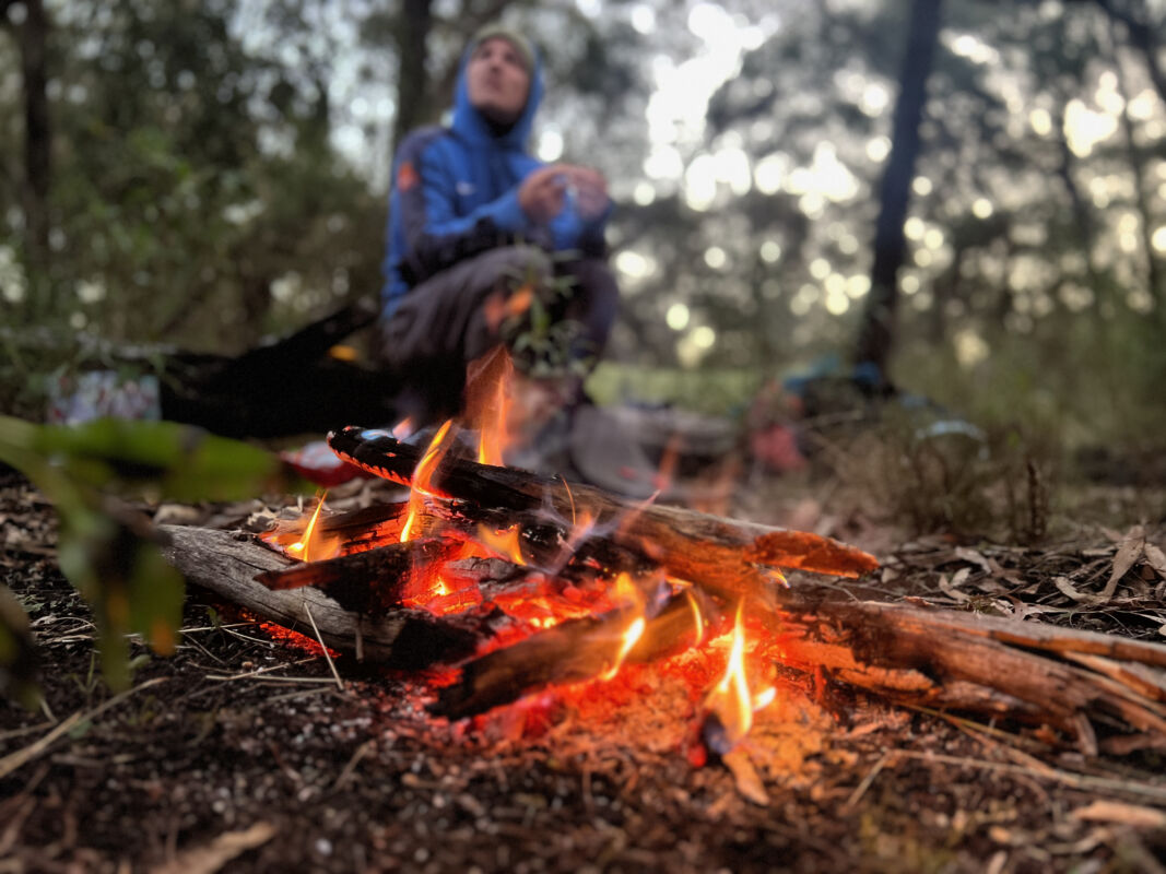 In the foreground is a small camp fire. This is what the picture is focused on. In the background a blurred climber is enjoying a cup of tea. The picture looks warm and inviting.