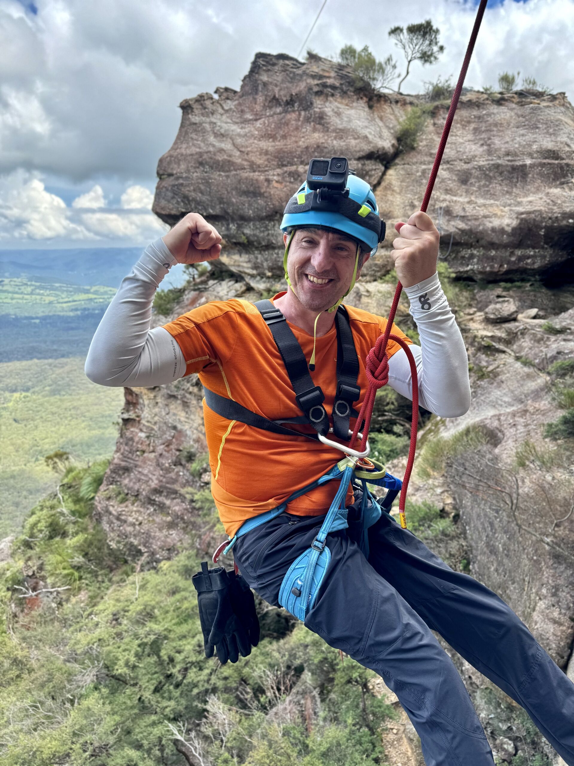 A smiling climber is shown in bright clothing, hanging in a rope. He is wearing a helmet and harness, as well as a chest harness. He is flexing the muscles in one arm while holding the rope he is suspended from in the other hand. He looks a long way off the ground. The climber is paraplegic and having an accessible adventure.