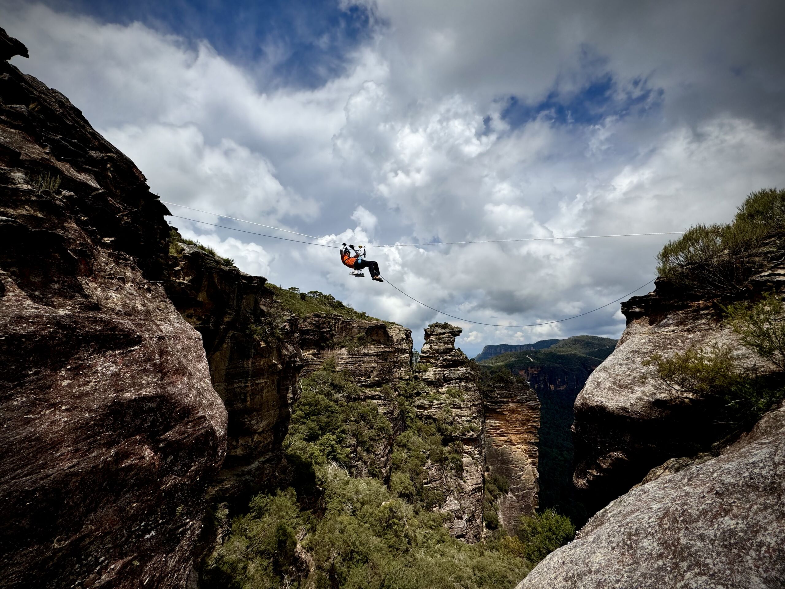 A climber is hanging from a Tyrolean Traverse. A rock feautre resembing a pigs head is in the background of the pic. Mike is a long way off the ground. The climber is paraplegic and having an accessible adventure.