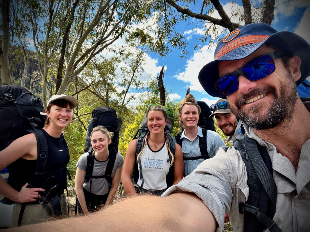 A group of six climbers stand taking a group selfie. They all look happy and clean. They are all wearing big packs and look ready to start walking in to their adventure. 