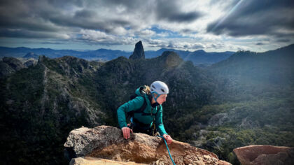 A Climber is pictured about to move onto a ledge. The background is spectacutlar with the sun rising on Belougery Spire. It looks very adventurous, which it is.